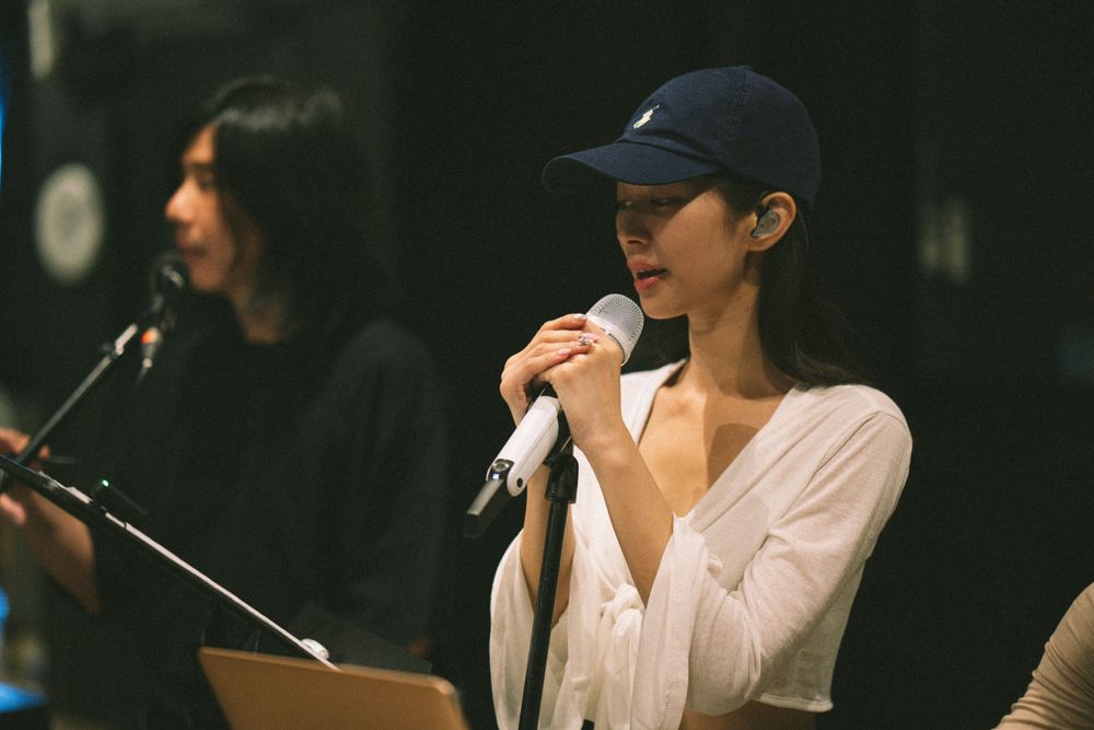 Vocalist in baseball cap and white shirt performs with microphone in dark studio setting.