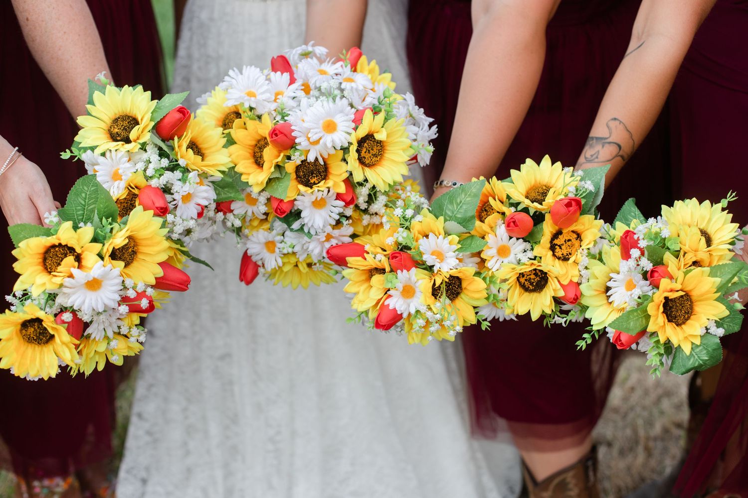 Close-up of colorful wedding bouquets featuring sunflowers and mixed flowers against burgundy dresses.
