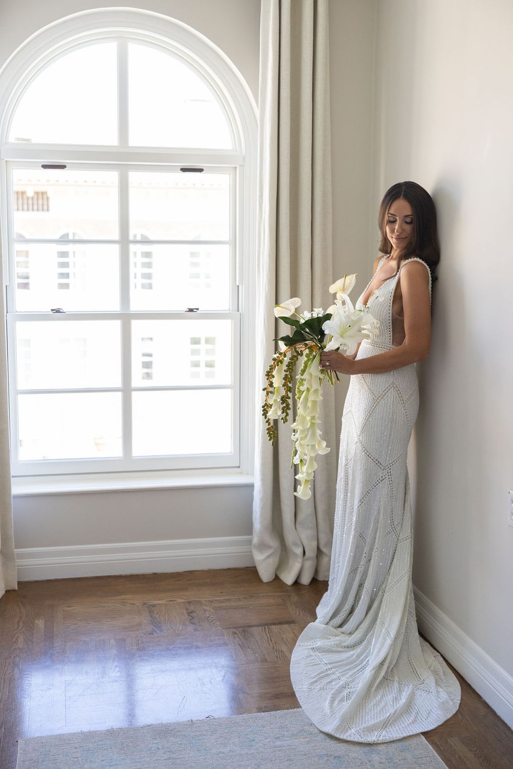 Woman in white wedding dress holding cascading bouquet of white flowers by arched window.