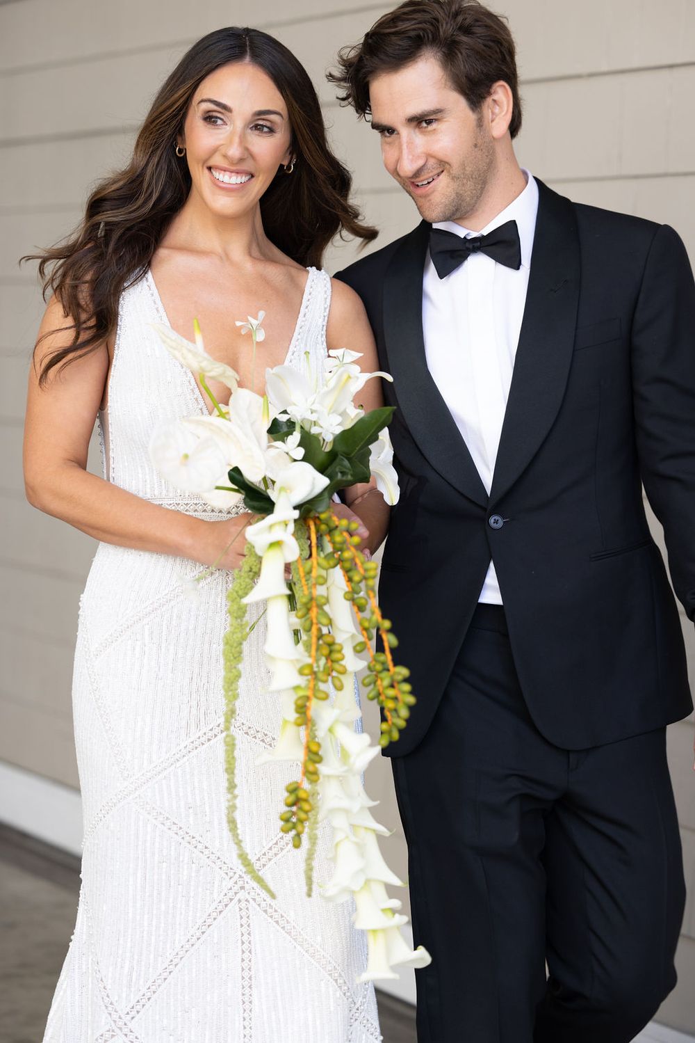Bride in white dress holding bouquet stands with groom in tuxedo, both smiling at camera.