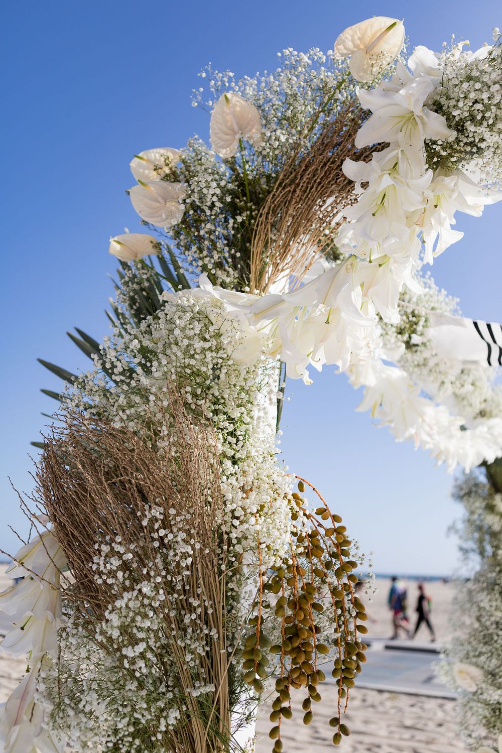 Floral arch with white flowers and greenery against blue sky, with people walking on beach in background.