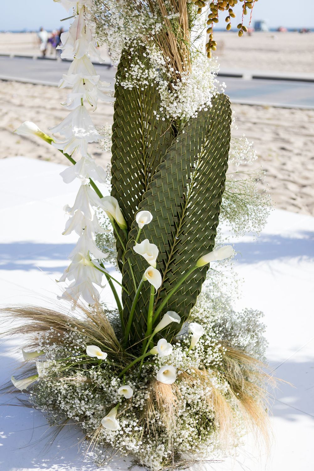 Tropical wedding floral arrangement with white flowers, palm leaves, and pampas grass on beach.