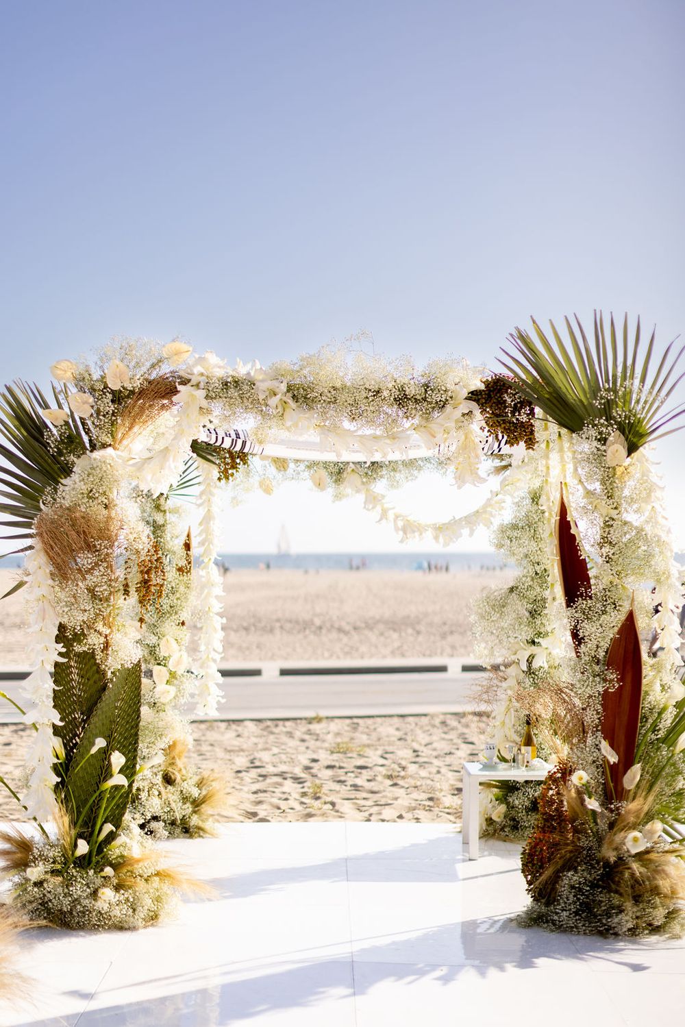 Rustic beach wedding arch decorated with white flowers, pampas grass, and palm leaves.