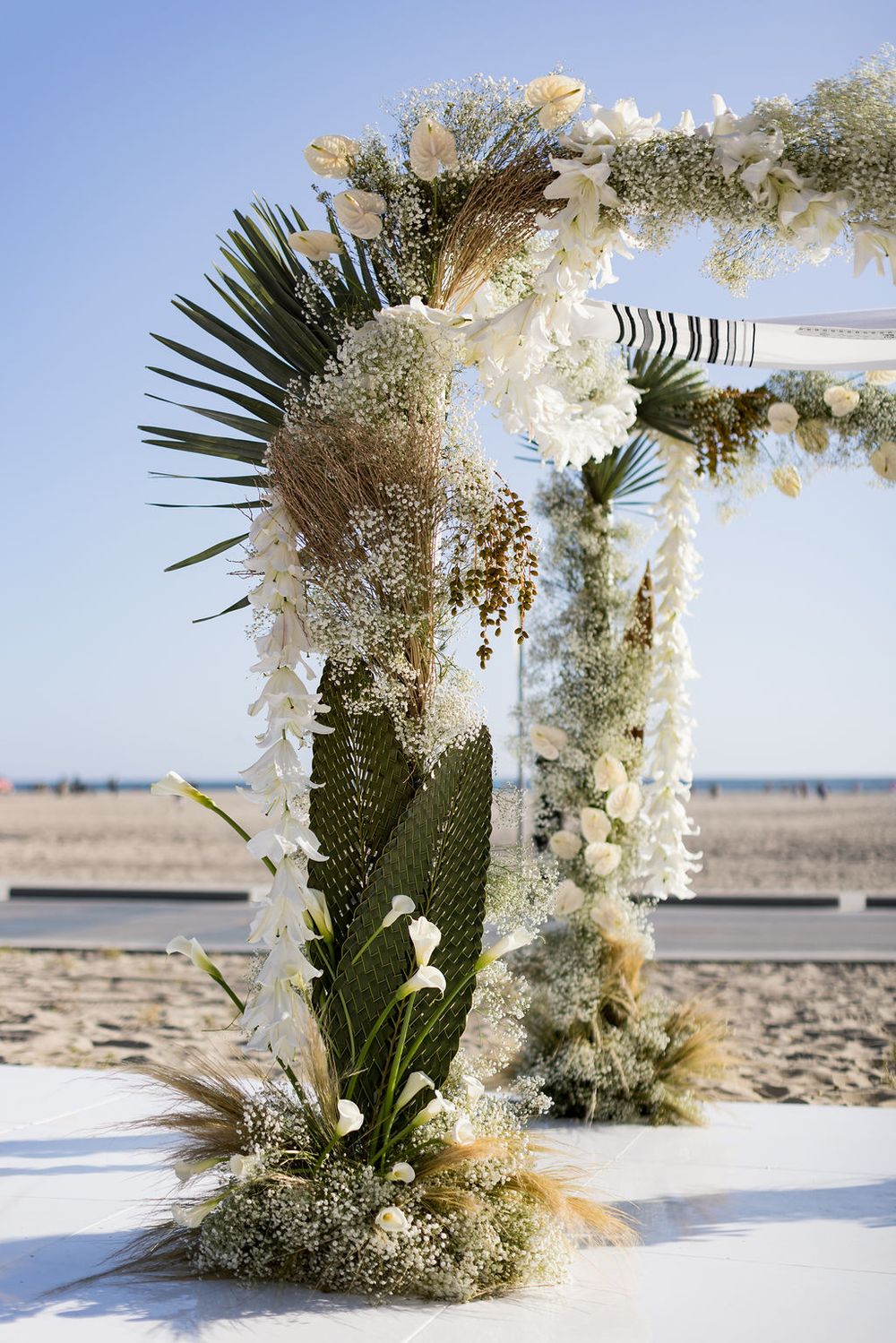 Beachside wedding arch with tropical flowers, pampas grass, and palm fronds against blue sky.