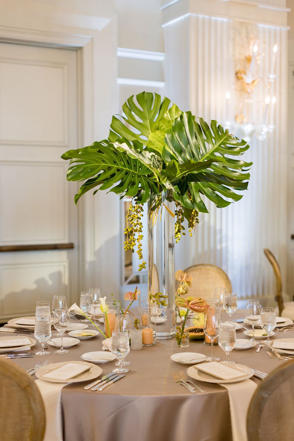 Elegant table setting with tropical centerpiece featuring large monstera leaves in a tall glass vase.