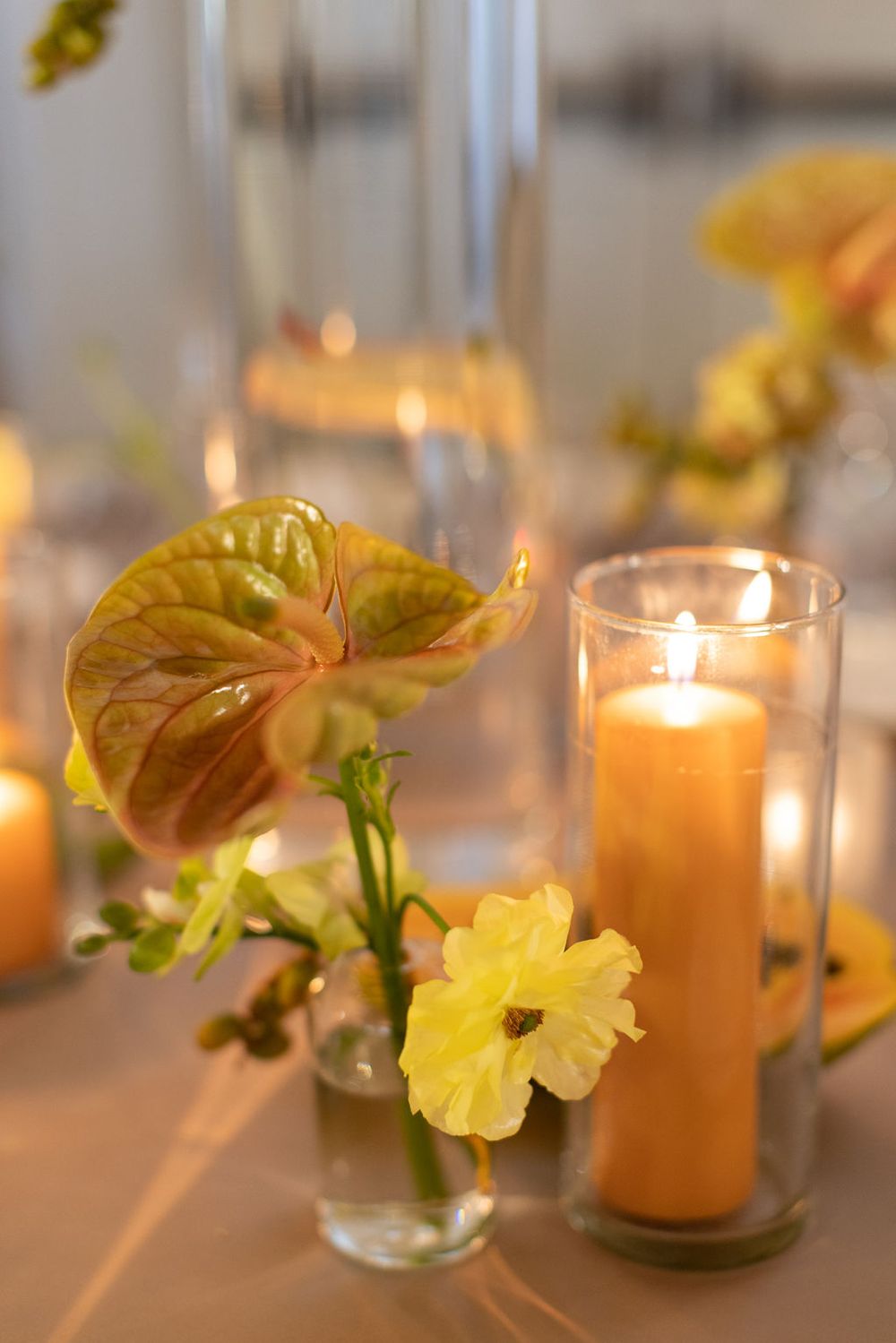 Romantic table decor with yellow flowers, anthurium leaves, and lit candles in glass holders.