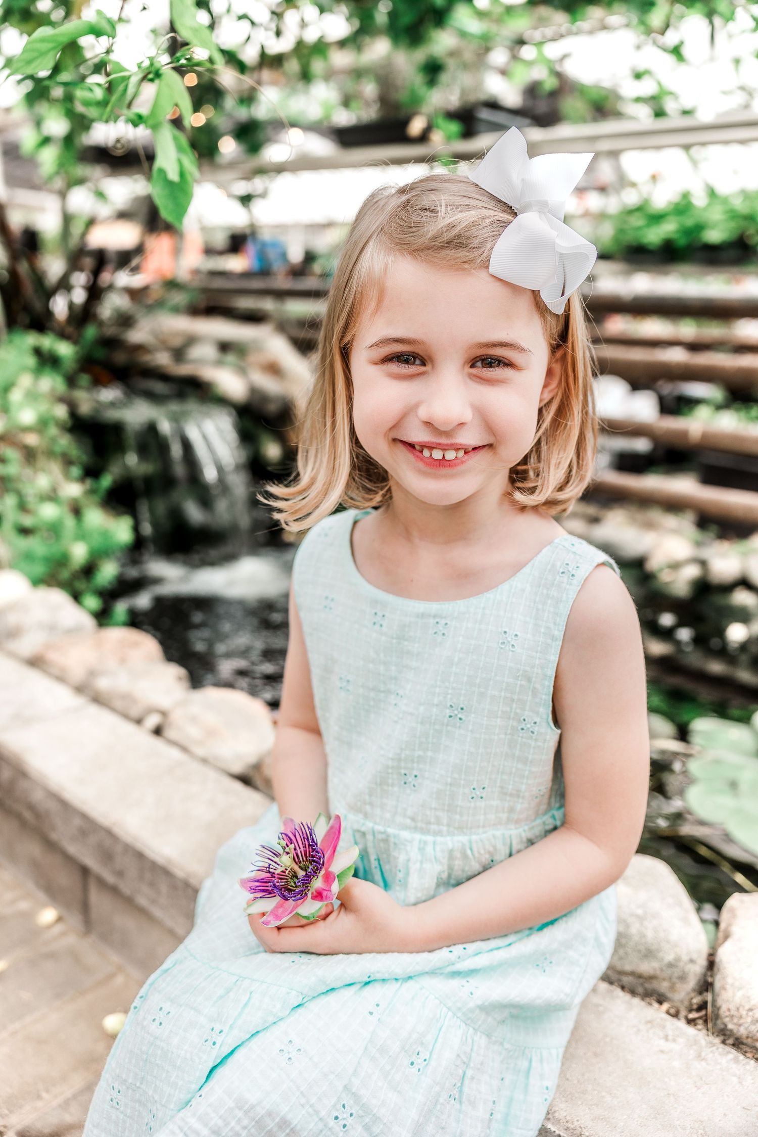 A figure in a mint dress holds a purple flower while sitting by a garden waterfall.
