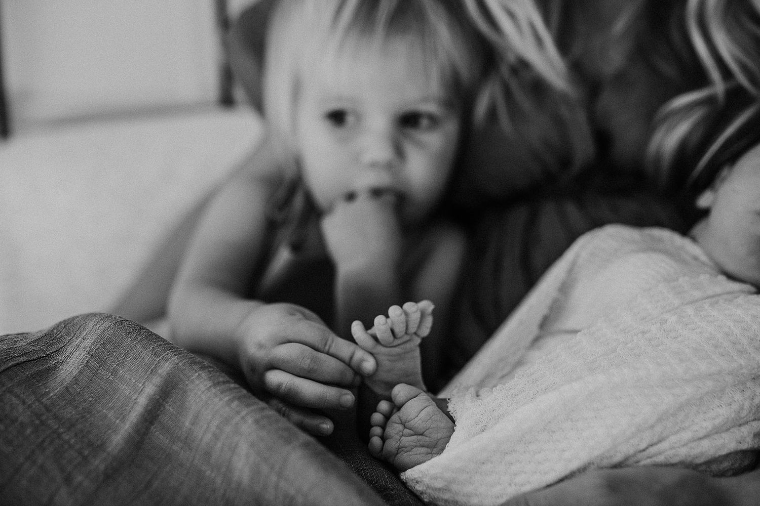 An older sister squeezes her newborn baby sister's toes during a newborn photoshoot.