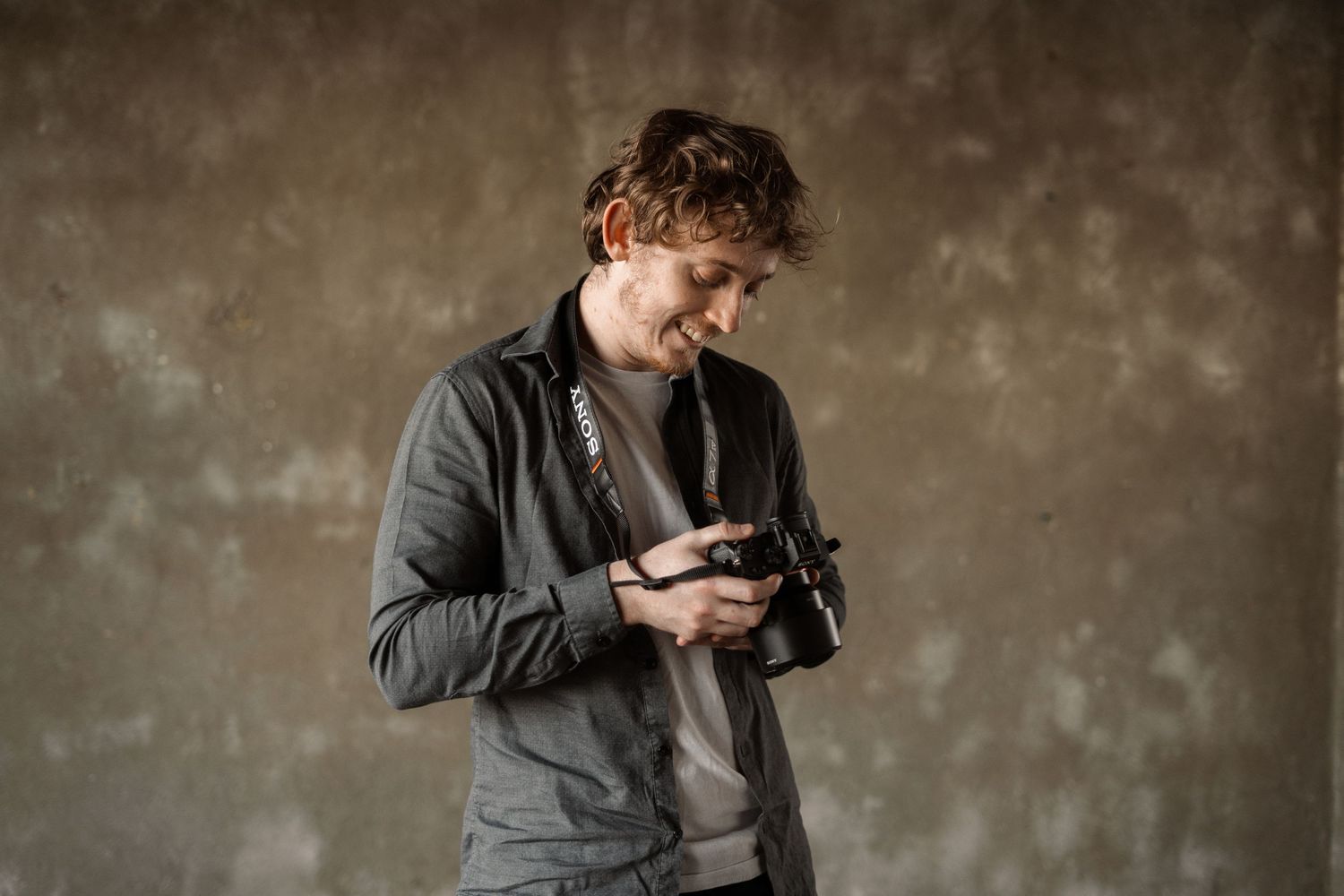 A photographer in casual attire examining a camera against a rustic wall background.