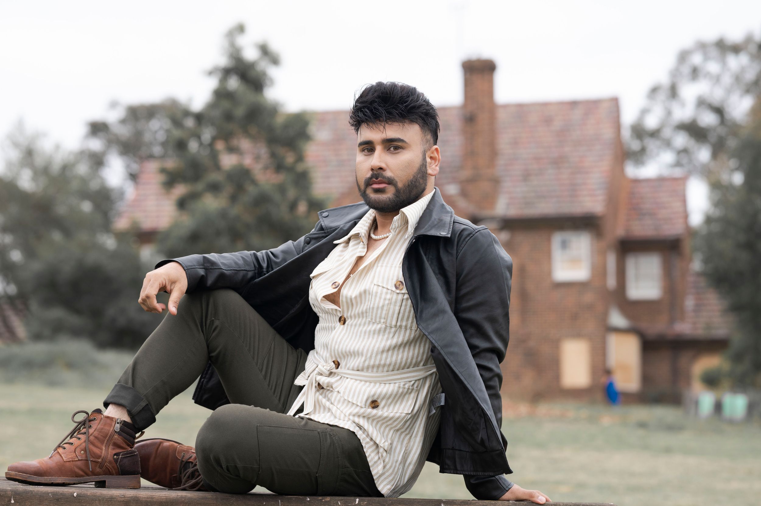Stylish bearded man in leather jacket and green pants sits casually outdoors near a brick building.