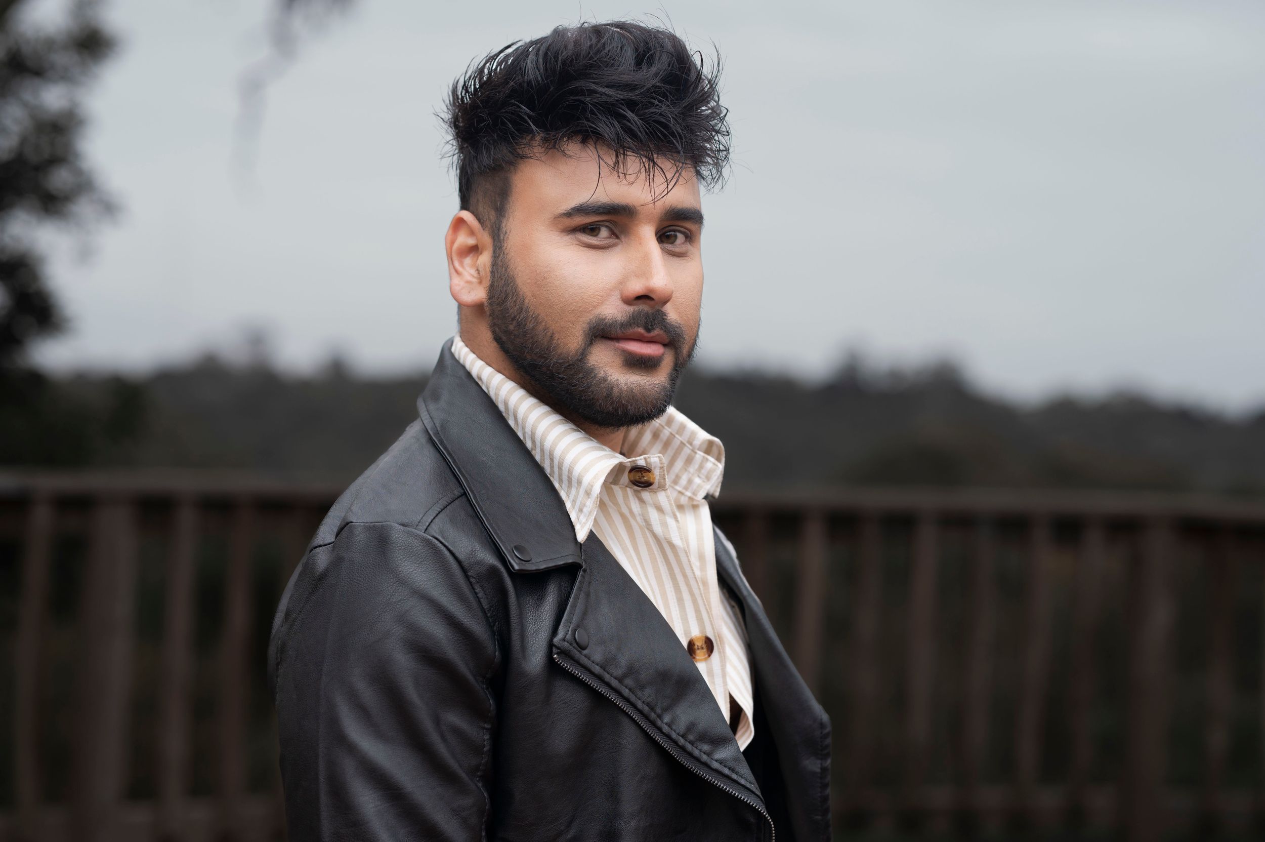 Young man with dark hair and beard wearing a leather jacket poses outdoors near a wooden fence.
