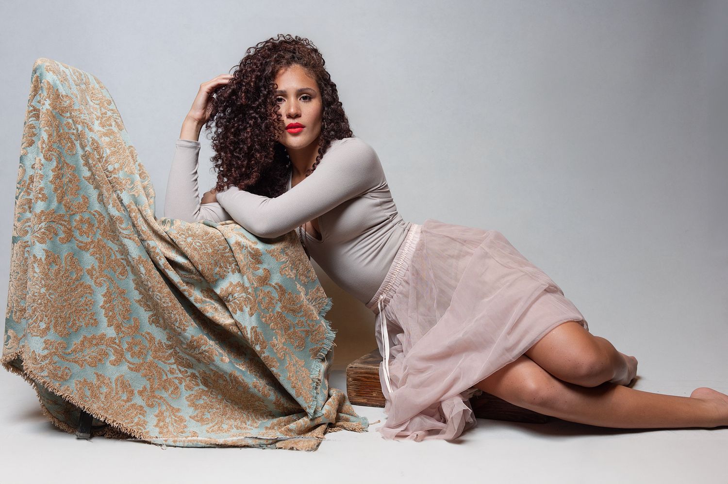 Woman with curly hair in a grey bodysuit and blush skirt posing with ornate fabric in a studio.