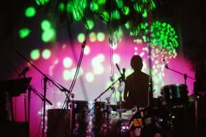 Silhouette of drummer performing on stage with dramatic green and pink lighting effects.