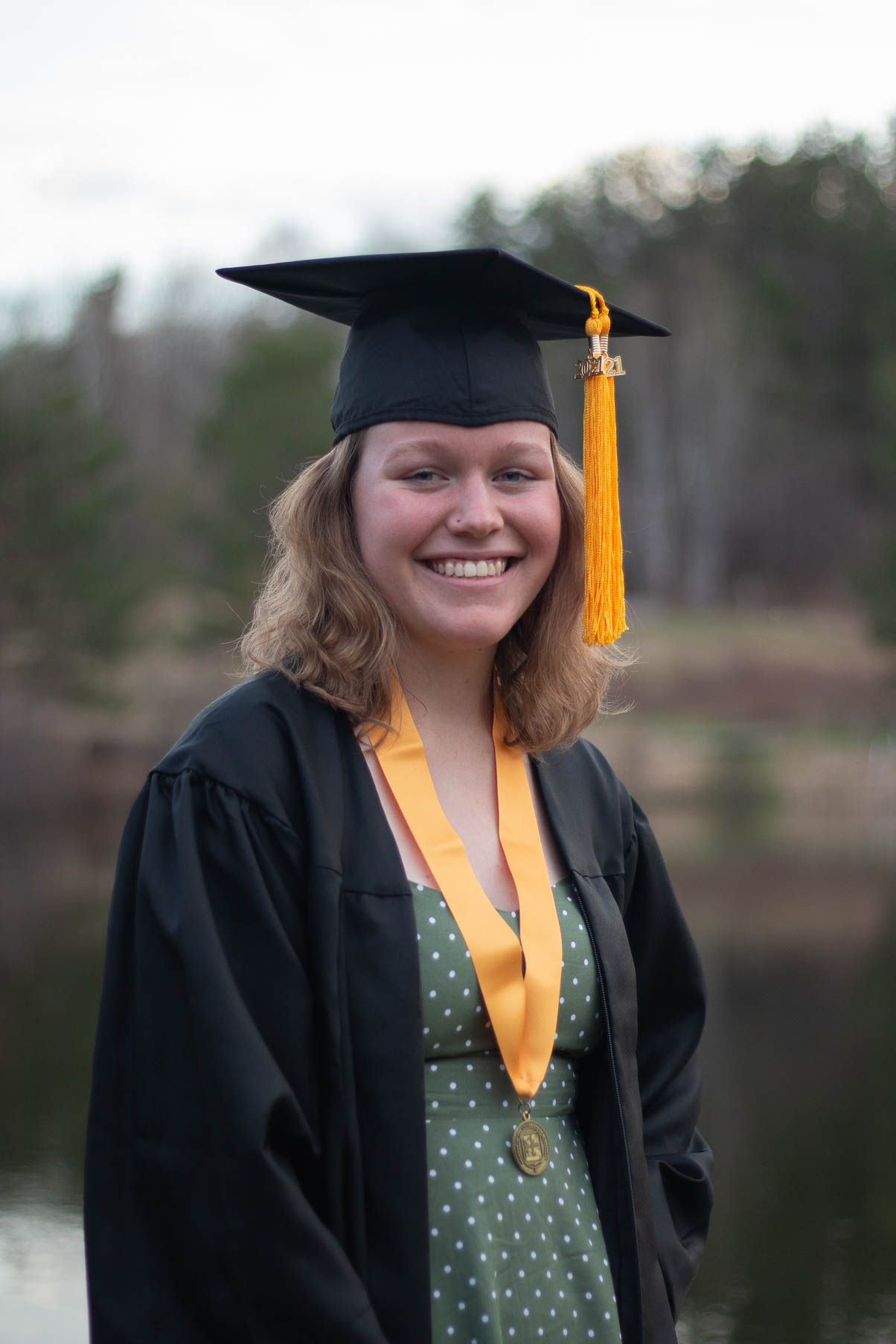 Graduate in cap and gown with yellow tassel posing by lake.