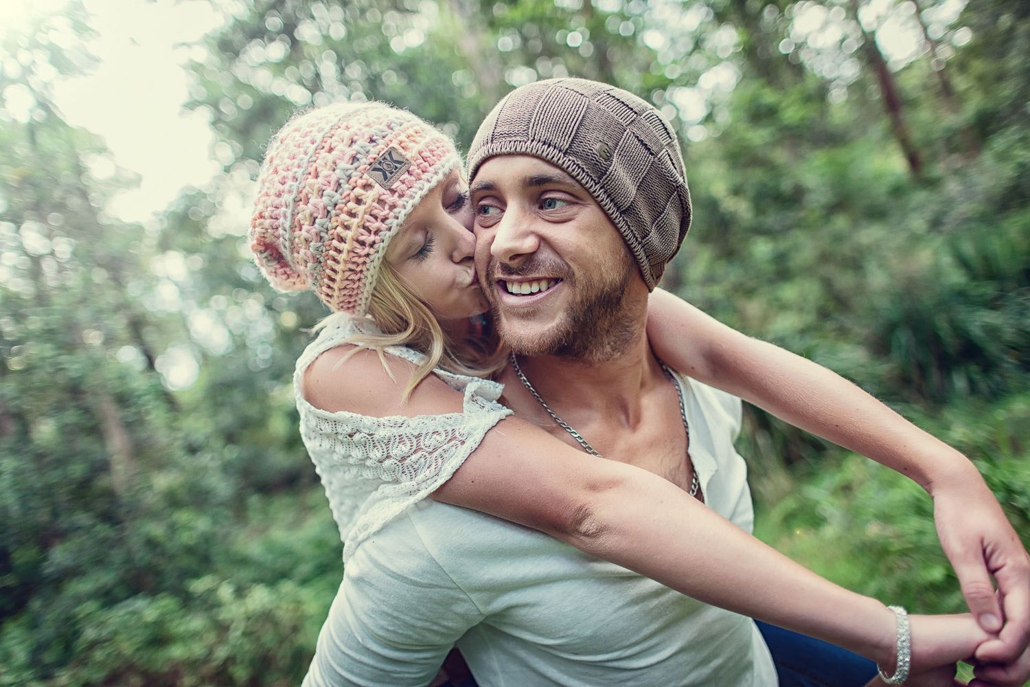 Young couple in beanies sharing a piggyback kiss outdoors in a lush green forest setting.