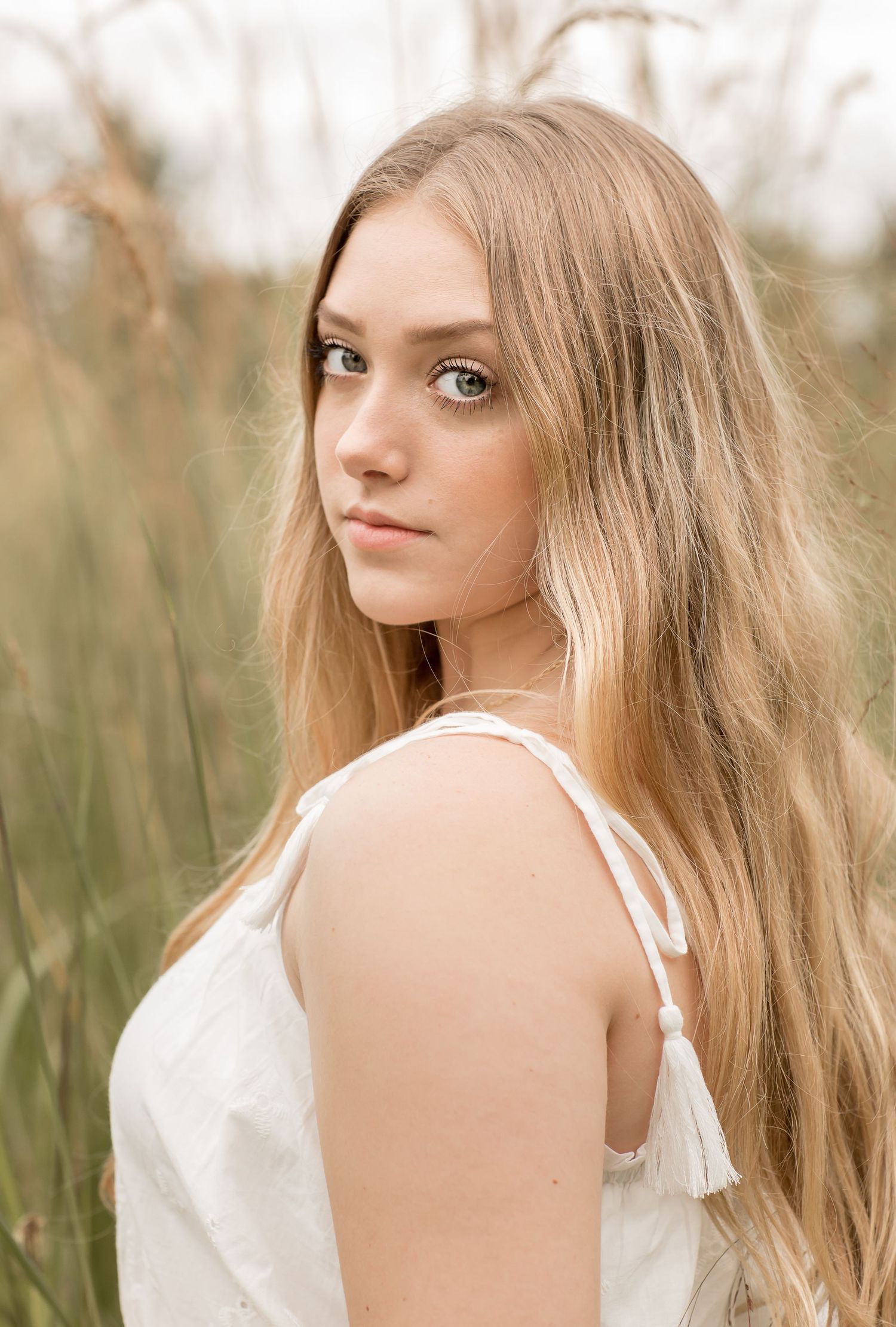 Young woman with long blonde hair in white dress poses outdoors among tall grass.