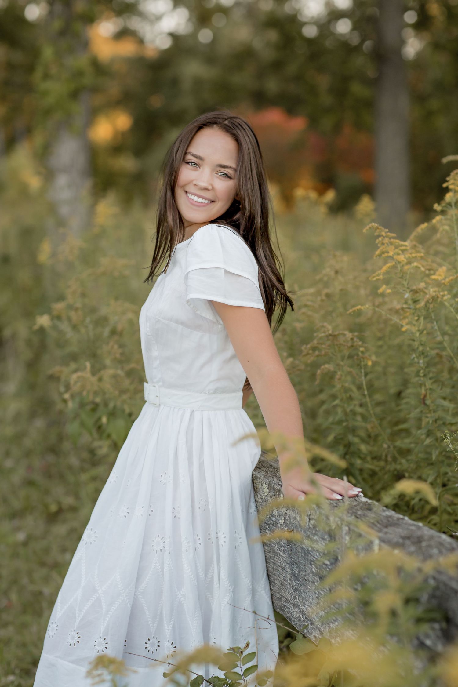 Smiling brunette teen in white eyelet dress leans on stone wall in autumn garden.