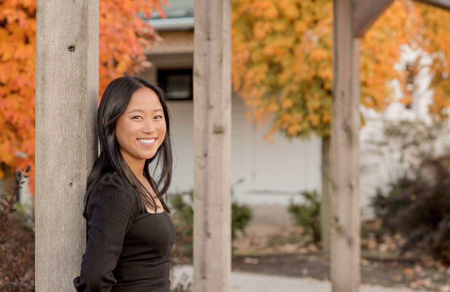 Smiling Asian woman in black top leaning against a wooden post with colorful autumn foliage behind her.