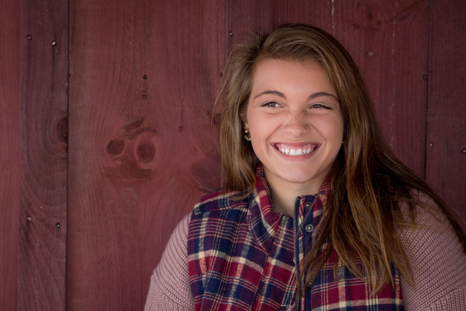 Smiling teen girl in plaid vest poses against rustic red barn wood background.