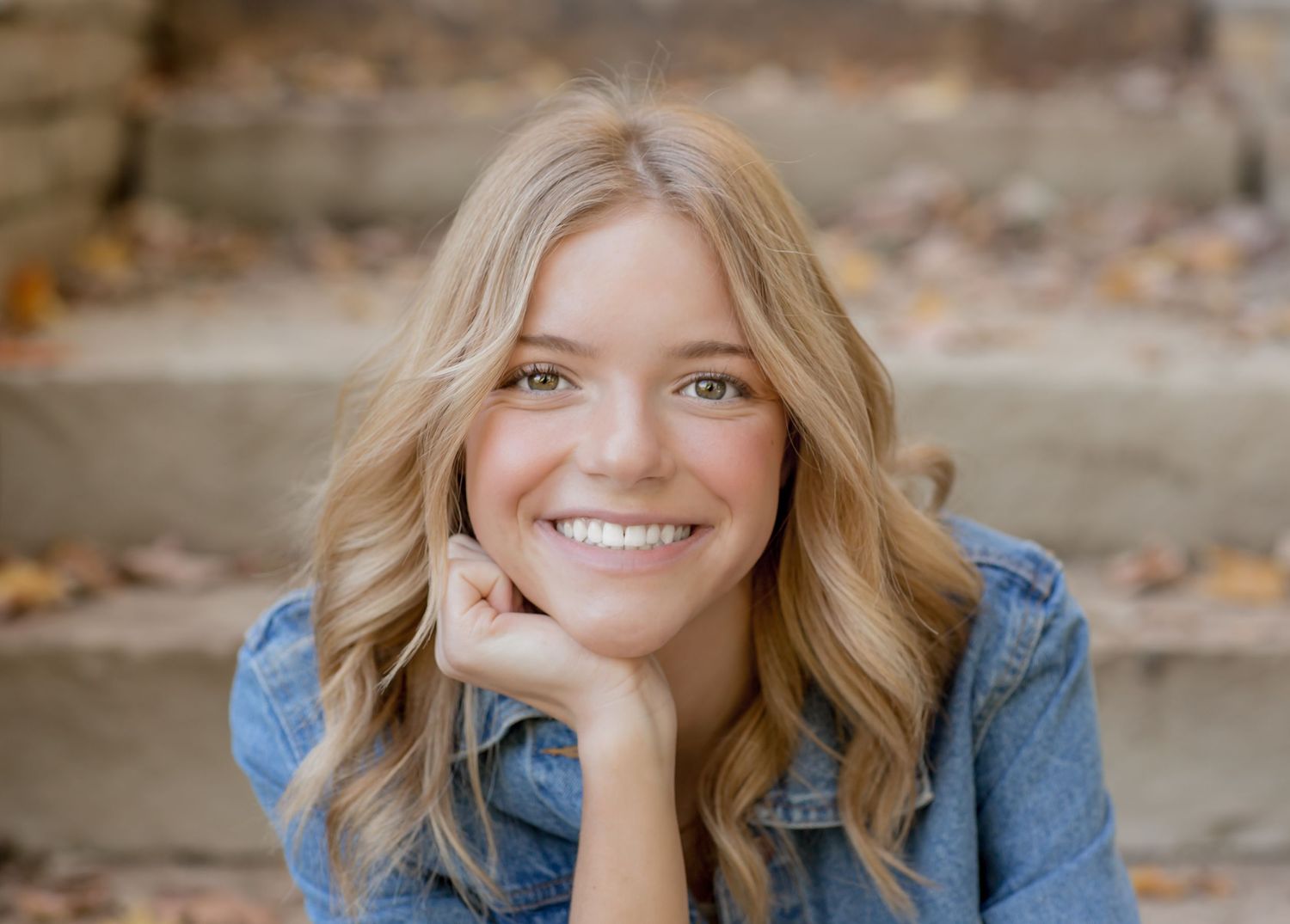 Smiling blonde teen with wavy hair poses on stone steps in denim jacket outdoors.