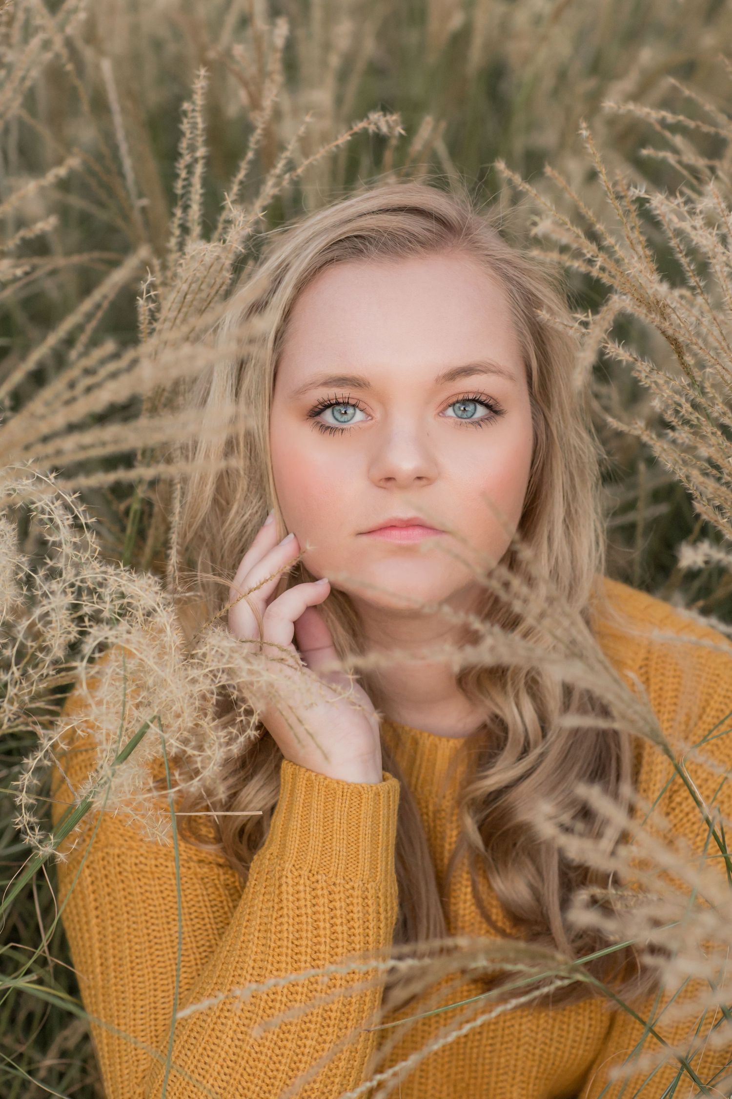 Blue-eyed blonde teenage girl in mustard yellow sweater posing among tall golden grass in a field.