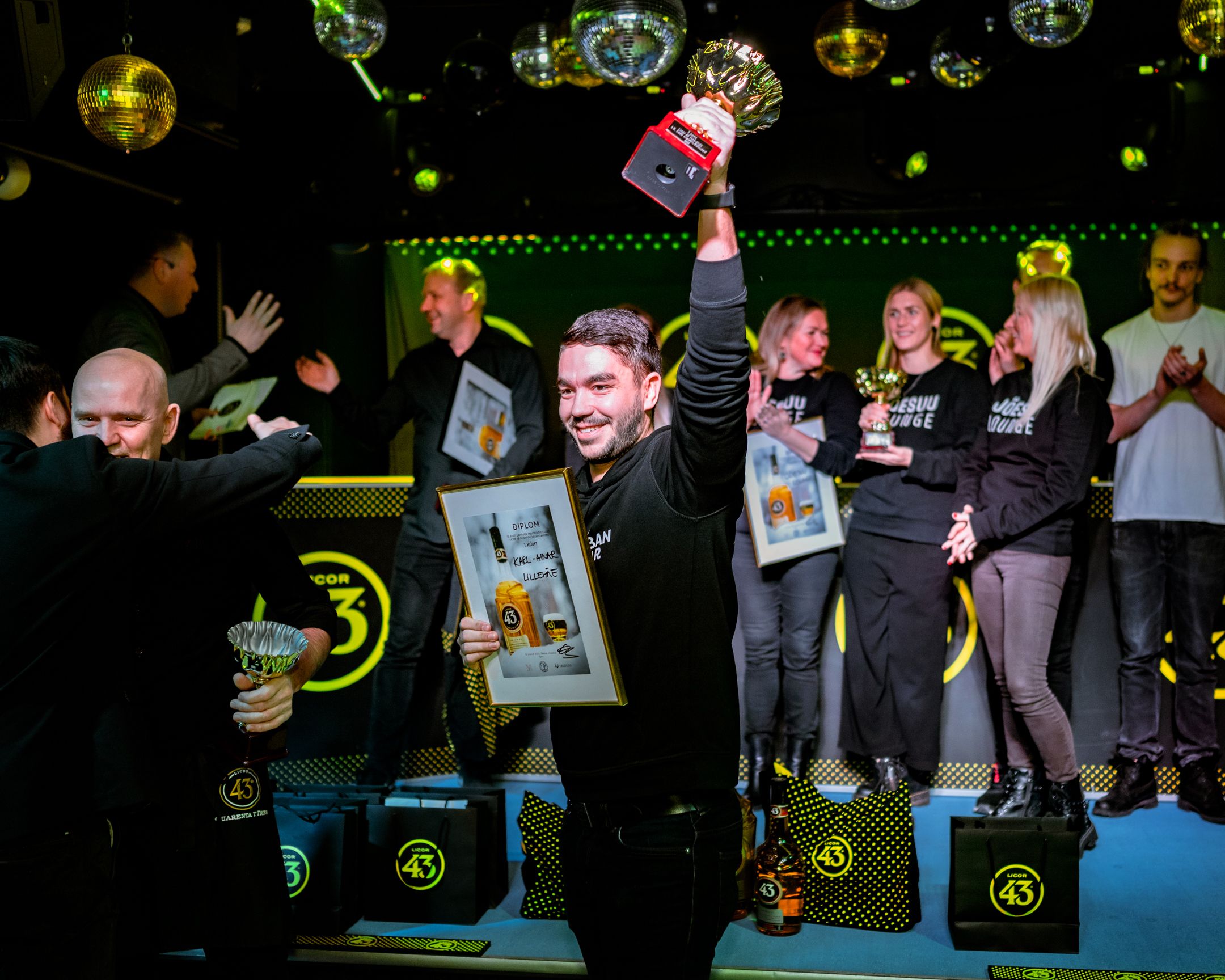 Winners celebrate on stage at an awards ceremony with trophies and framed certificates under colorful disco lights.