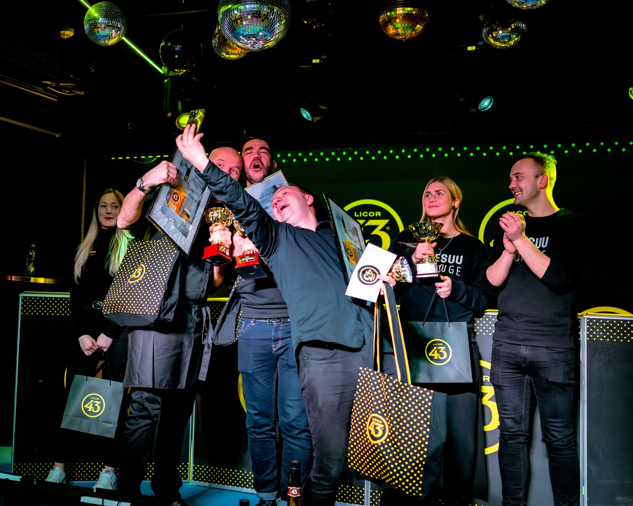 Winners celebrate enthusiastically on stage holding up trophies and awards at a nightclub event.