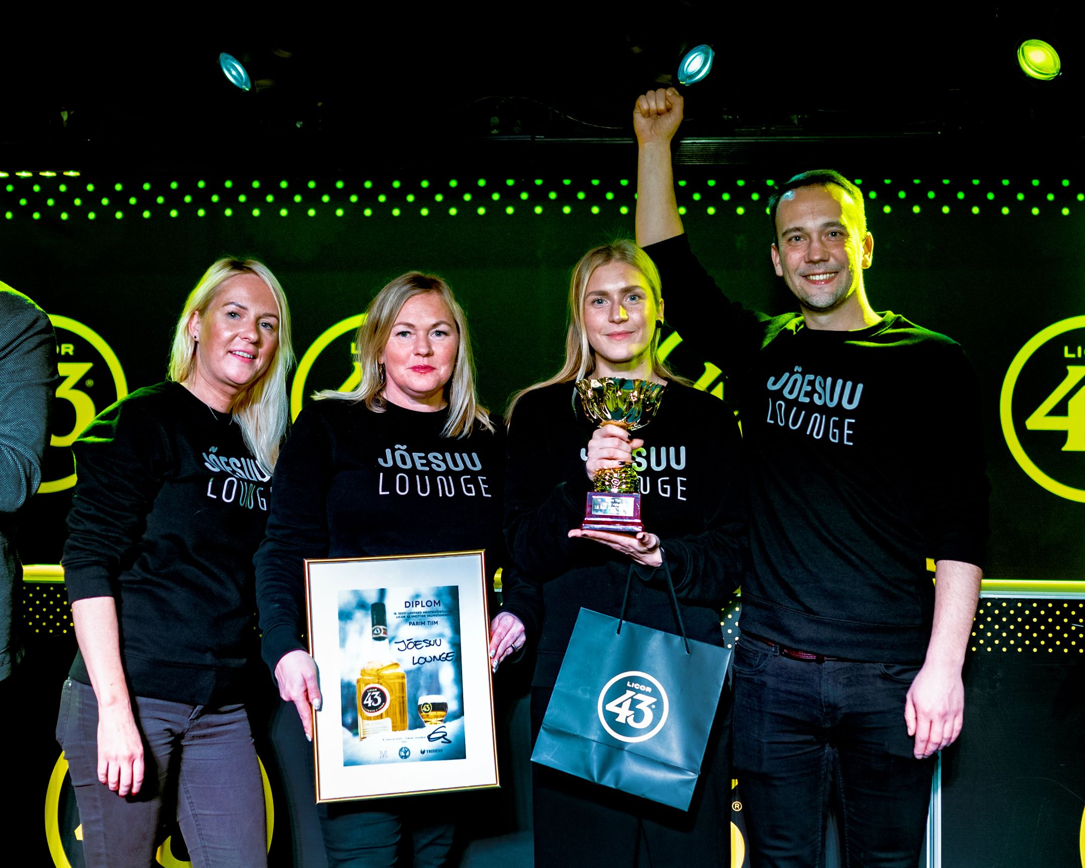 Award winners in matching black shirts pose with trophies and certificates on stage.