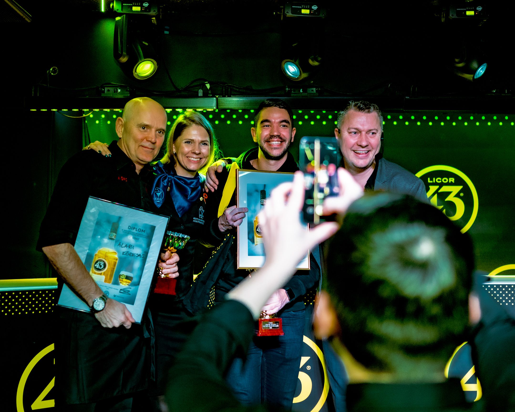 Group poses with awards and certificates under colorful stage lighting at celebration event.