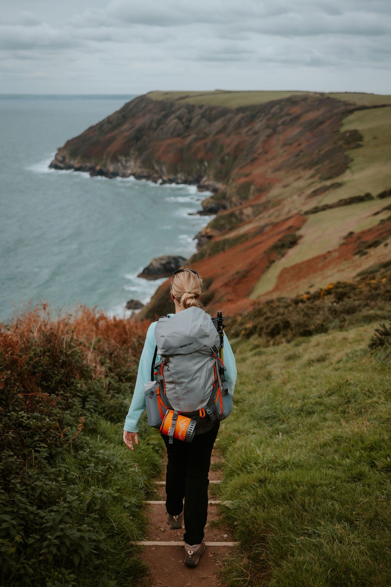 Hiker on coastal path with dramatic red cliffs and ocean views.