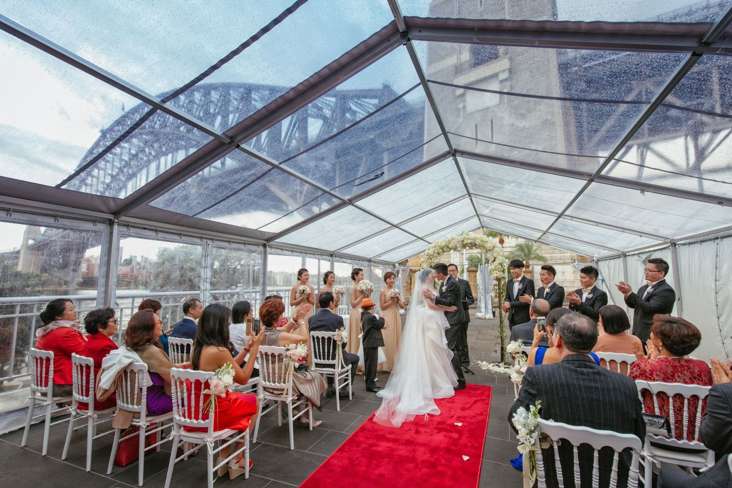 Wedding ceremony under clear roof marquee with Sydney Harbour Bridge backdrop, red carpet aisle and white chairs.