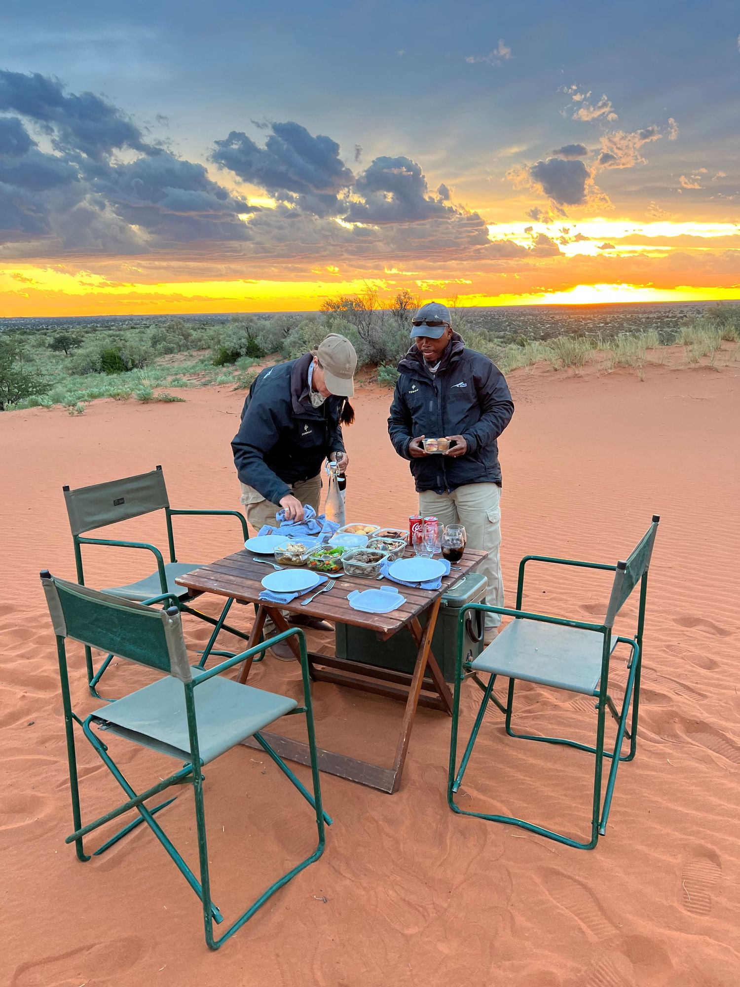 Outdoor dining setup with chairs and tables arranged on red desert sand during sunset.