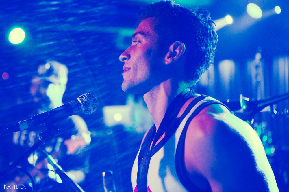 Close-up of a young male vocalist singing into a microphone under blue concert lighting.