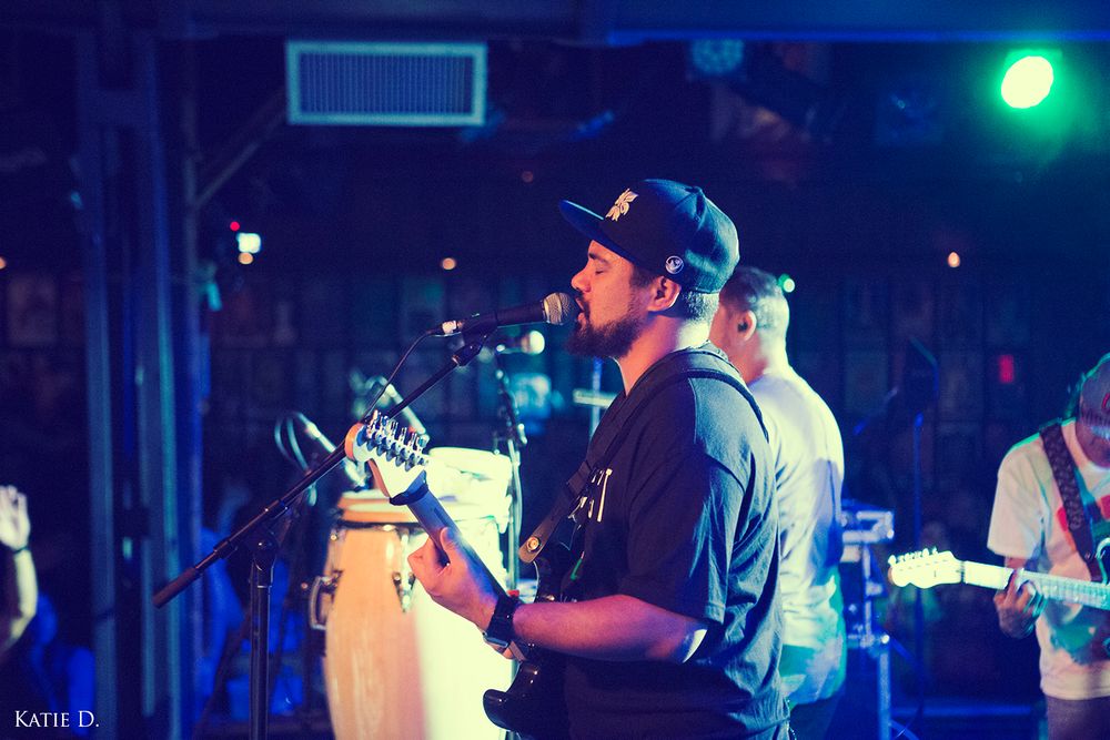 Guitarist singing into microphone on stage with blue lights and conga drums at live concert.