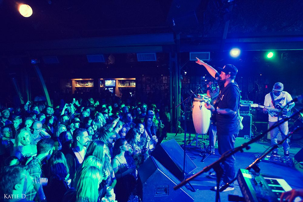 Band performing on stage to large crowd at energetic night concert with blue lighting.