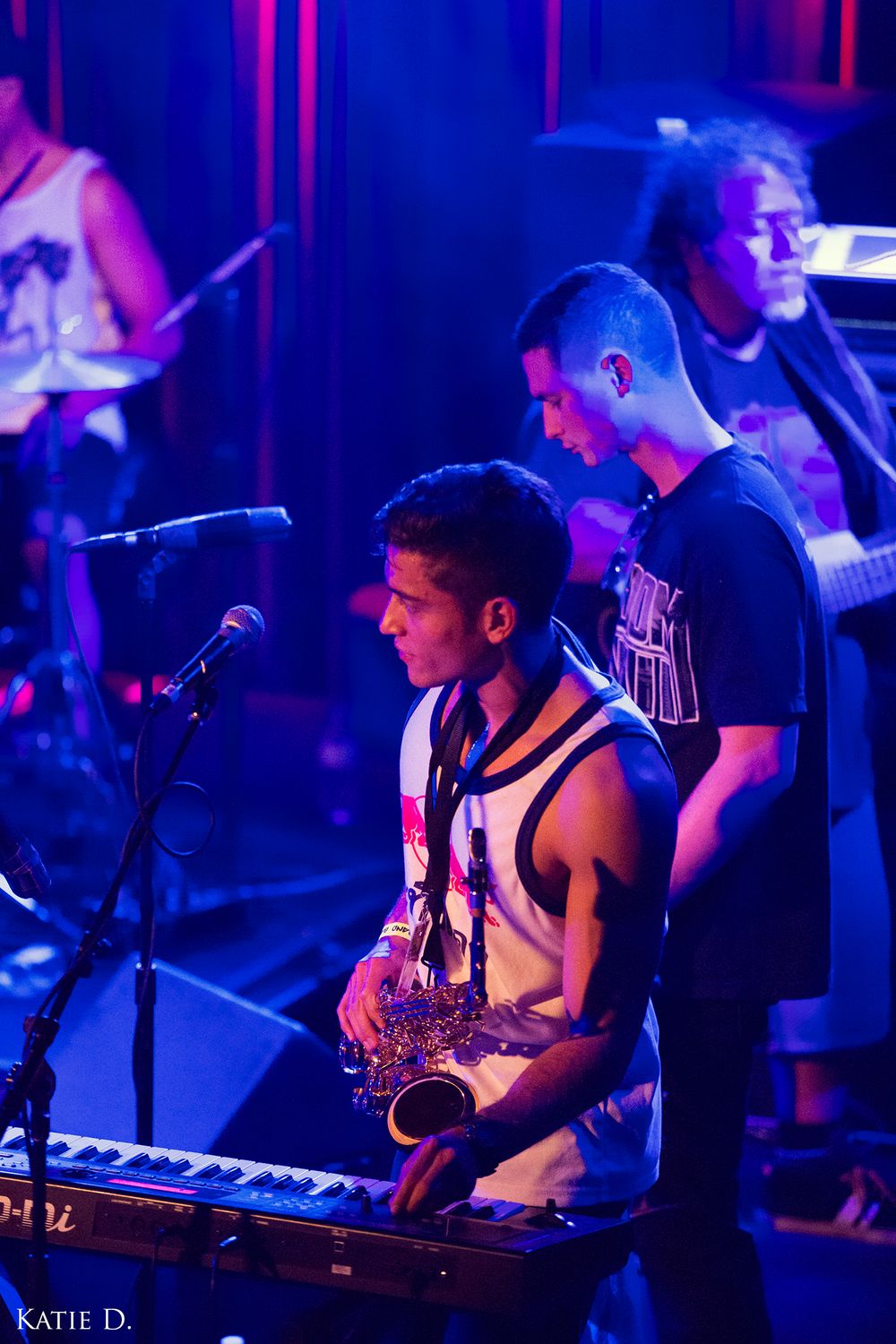Saxophone player performing on stage with keyboard and band members in purple concert lighting.