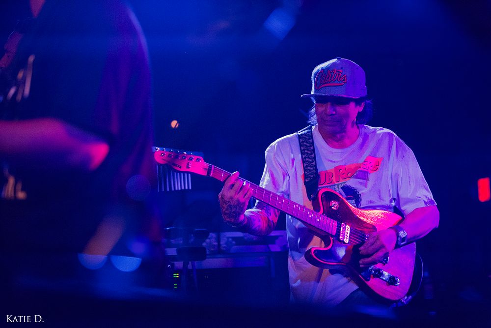 Guitarist in snapback cap playing electric guitar under blue stage lighting at concert.