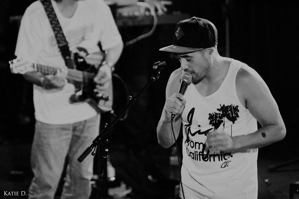 Singer in snapback hat and tank top performing into microphone with guitarist in background.
