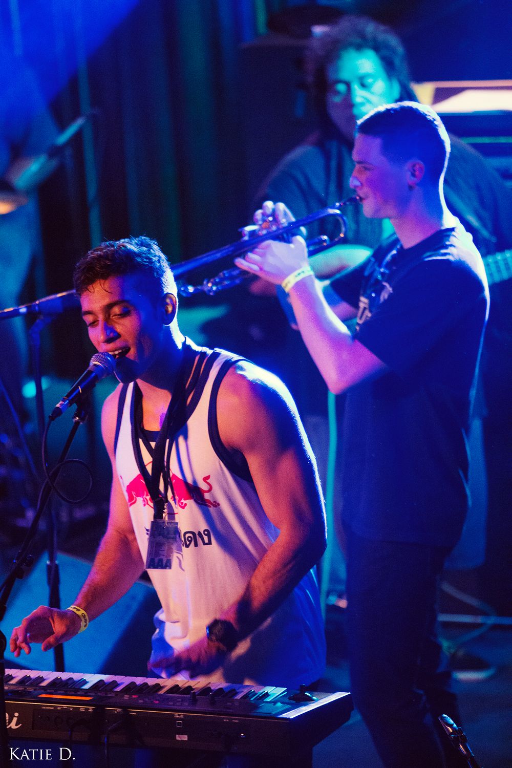 Keyboard player sings into microphone while trumpet player performs behind him on a colorful lit stage.