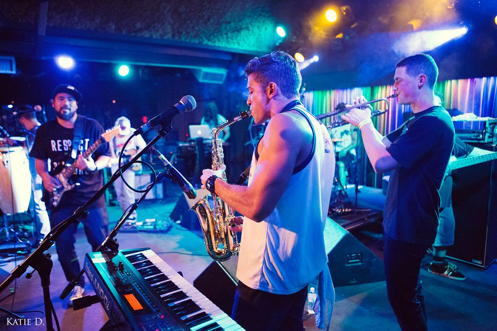Live band performance featuring saxophone, keyboard, and trumpet players under colorful stage lights.