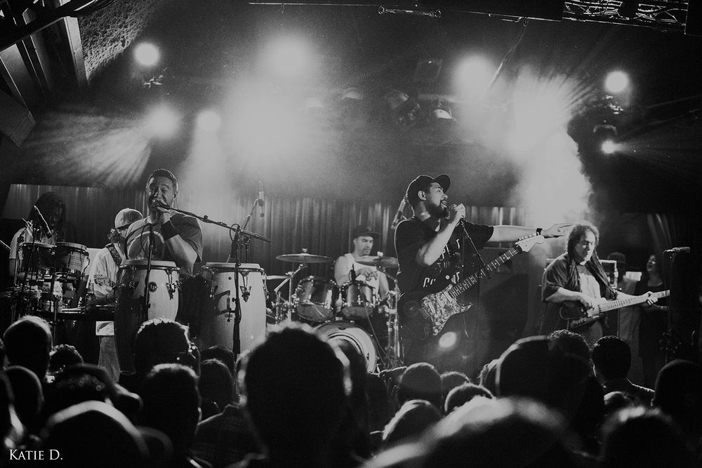 Black and white wide shot of a full band performing live on a lit stage for a crowd.