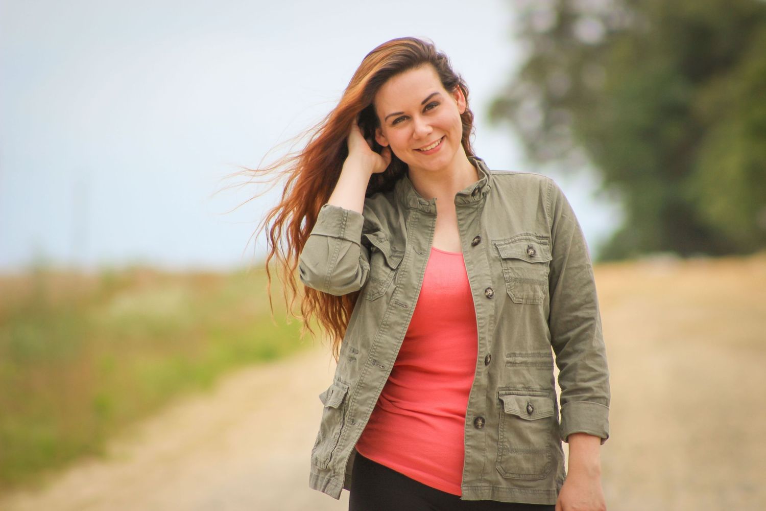A woman in a green utility jacket and coral top poses outdoors on a rural path with wind-blown hair.