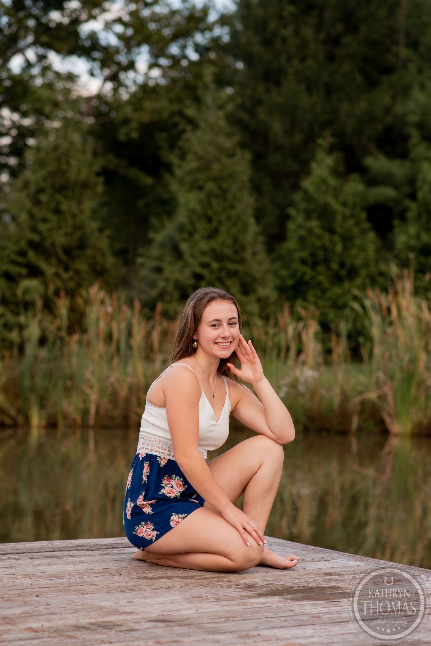 Summer portrait of someone in floral shorts sitting cross-legged on a lakeside dock surrounded by evergreens.