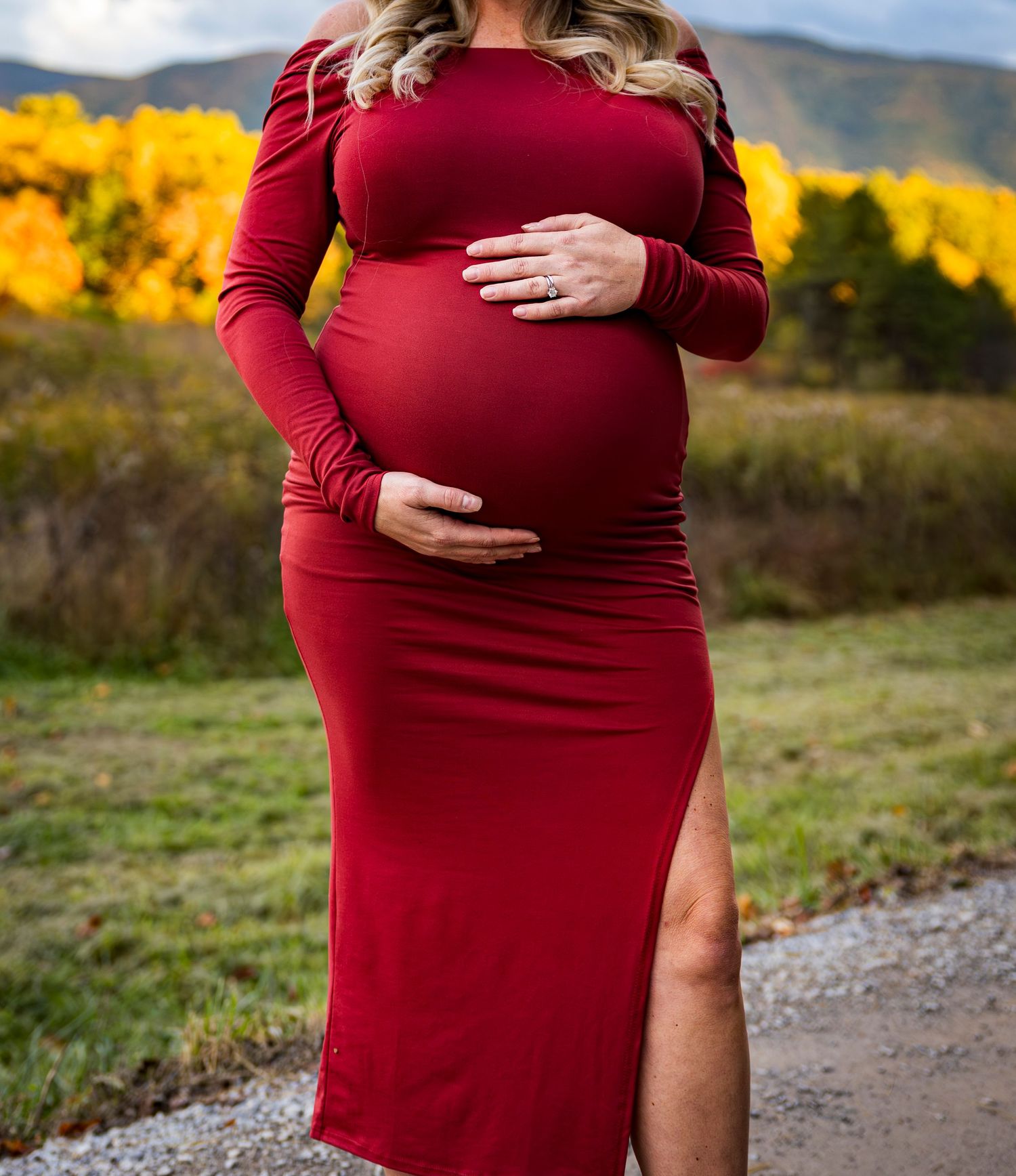 Close-up of pregnant belly in red dress with hands cradling bump against autumn landscape.
