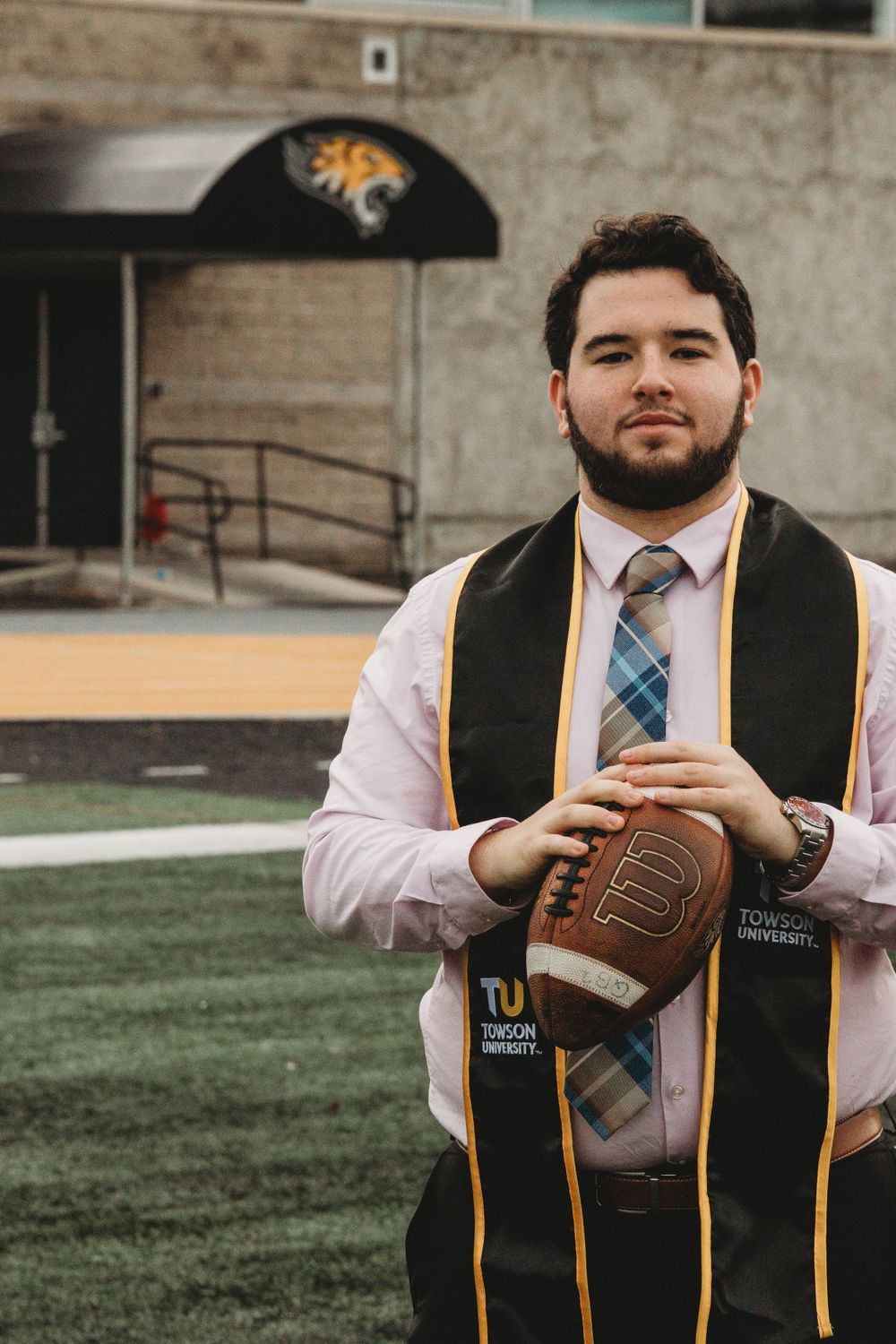 Graduate in academic regalia posing with football on campus lawn.