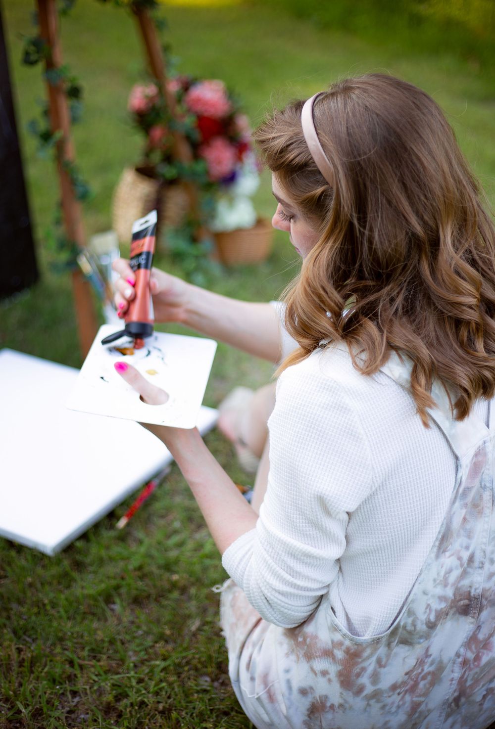 Artist in white sweater painting in an outdoor garden setting with art supplies and American flag.