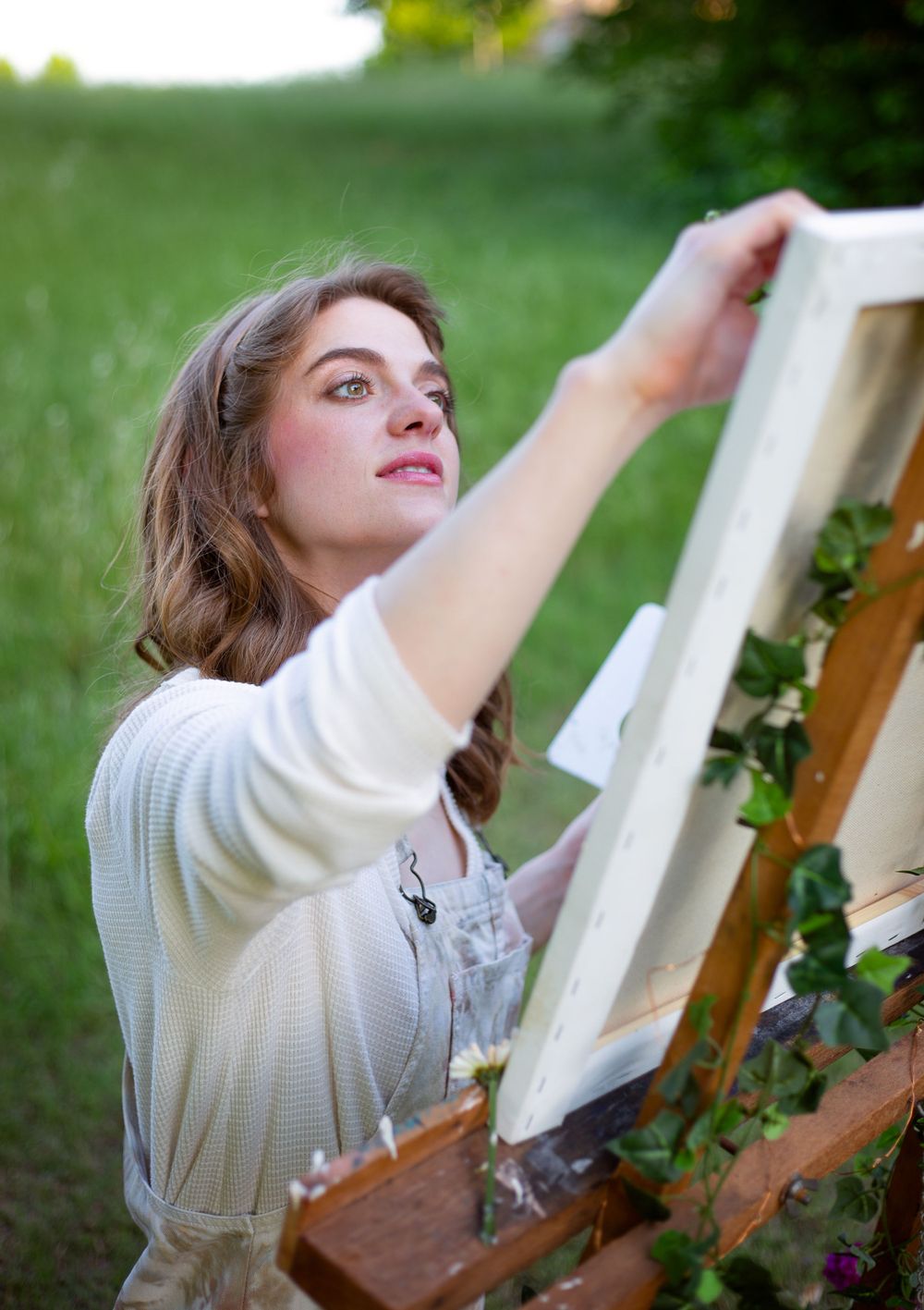 Artist working at an easel outdoors in a white sweater and overalls.