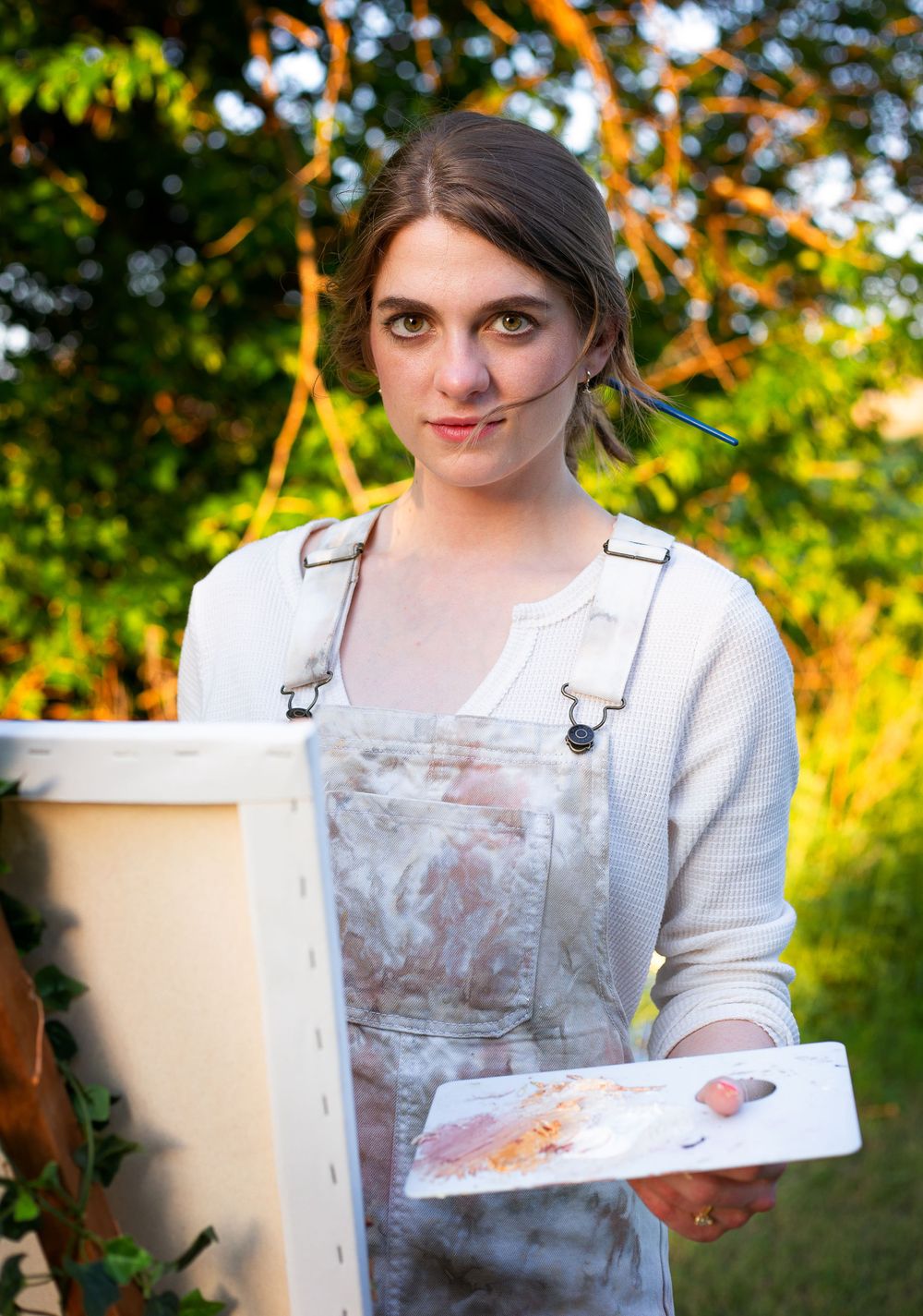 Artist in white sweater and paint-stained overalls holding art supplies in garden setting.