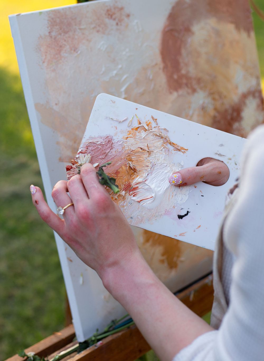 Paint-stained hands holding an artist palette against a canvas outdoors.