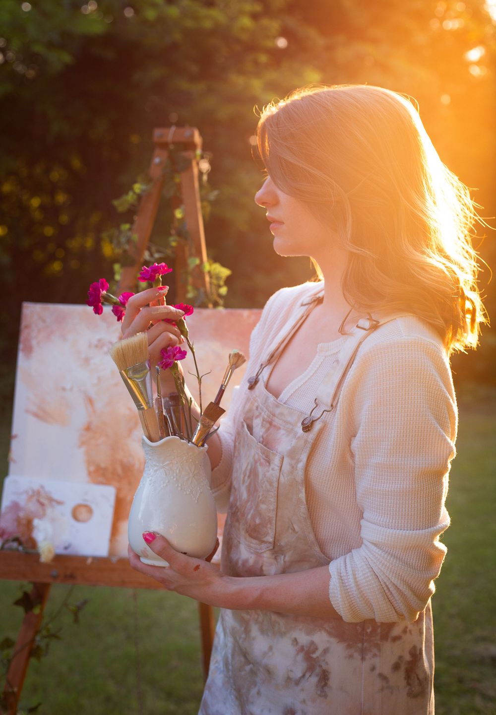 Artist in white dress holds flowers against golden sunset light while painting outdoors.